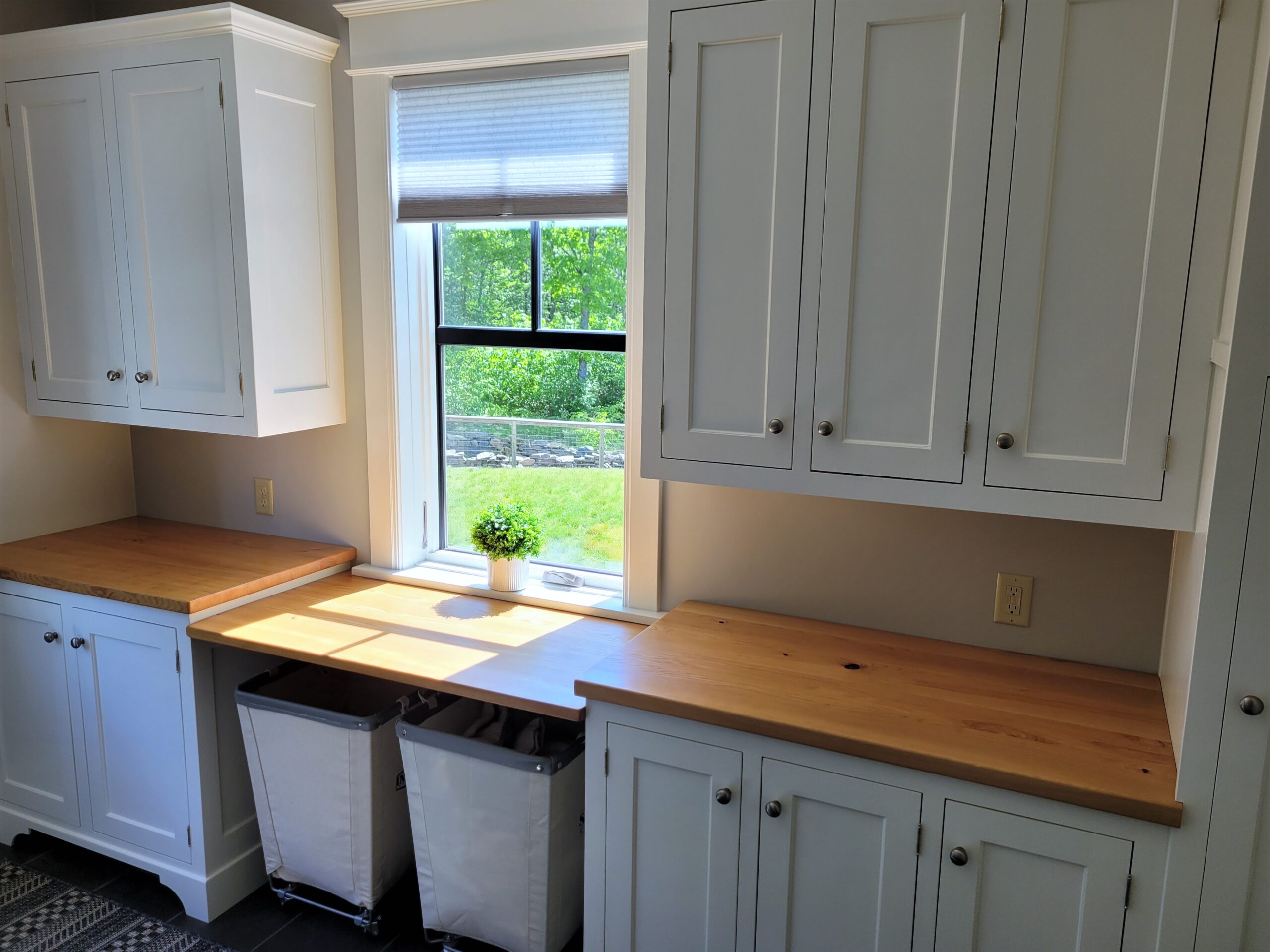 Photo of laundry room with white upper and lower cabinets separated by a pine counter that functions as a folding station. Two canvas wheeled laundry carts are parked under the folding counter.