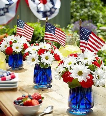 Three blue glass vases filled with daisies, red carnations, and small American flags.