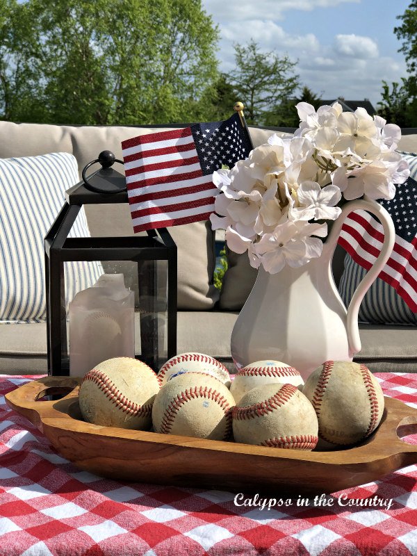 A shallow wooden bowl filled with old baseballs sits next to a white pitcher with white hydrangeas and small American flags in it.