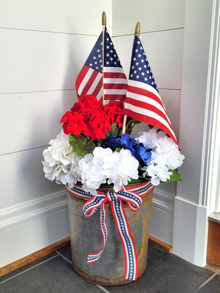 Memorial-Day-Arrangement | The Granite Acorn Rusty sap bucket filled with white and blue hydrangea red geraniums and two american flags The bucket is tied with a red white and blue ribbon