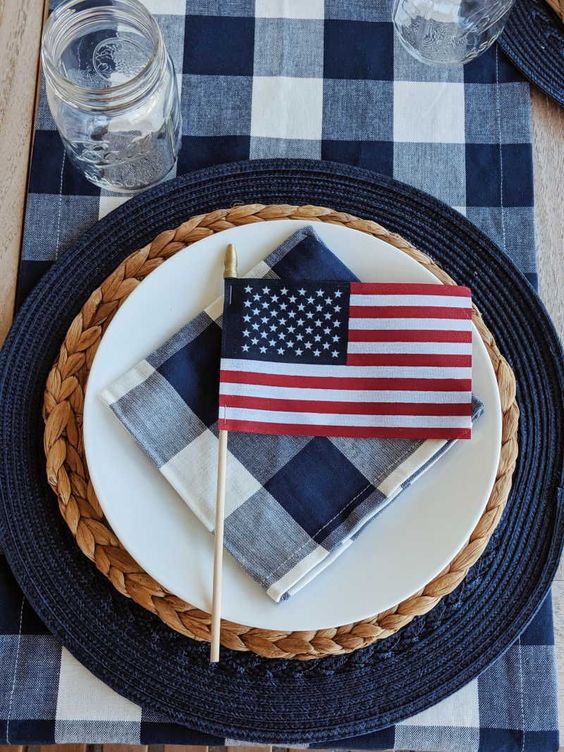 A place setting with a dark blue round placemat, hyacinth charger plate, white dinner plate, blue and white checked napkin and small American flag.