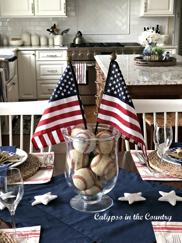 Footed glass vase filled with old baseballs and two small American flags on a dining table.