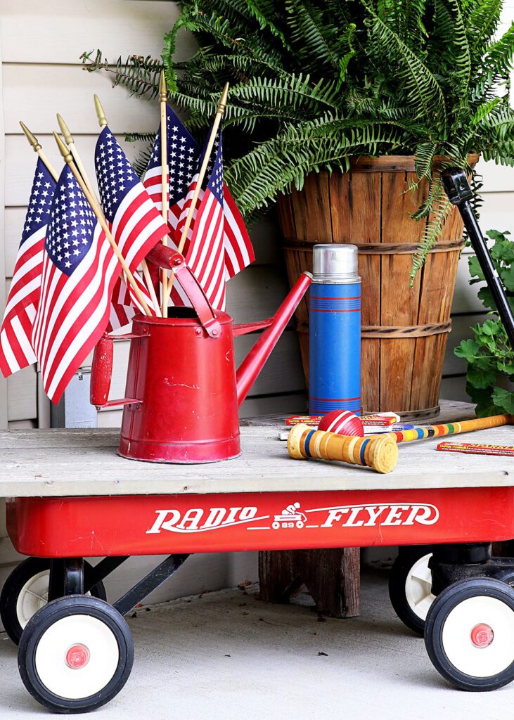 A child's red wagon topped with a piece of wood. A red watering can filled with small American flags and a blue thermos sit on top of the wood. The wagon is parked by a potted fern on a porch.