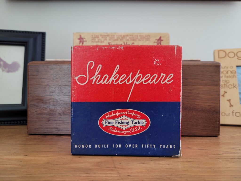 Close up of a vintage fishing reel box. The box is red and blue and says Shakespeare on it.