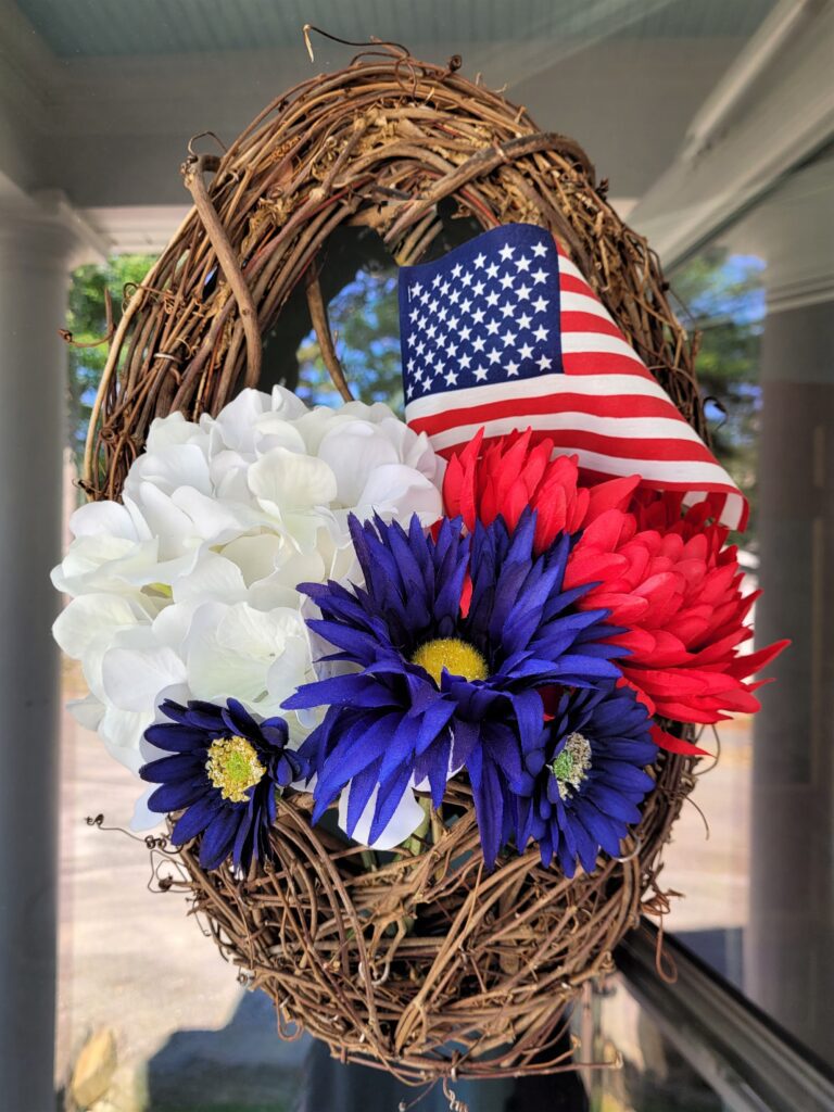 Wicker wreath basket with a white hydrangea, blue daisies, red mum, and small American flag