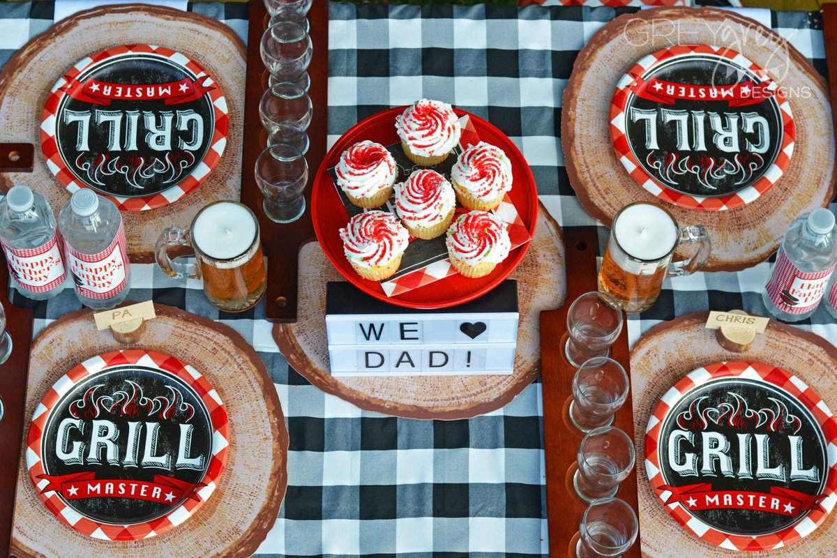 Picnic table with red and white paper plates, faux wood chargers, beer mug candles, a black and white checked tablecloth, water bottles that say Happy Father's Day, and a red cake stand with cupcakes as the centerpiece.