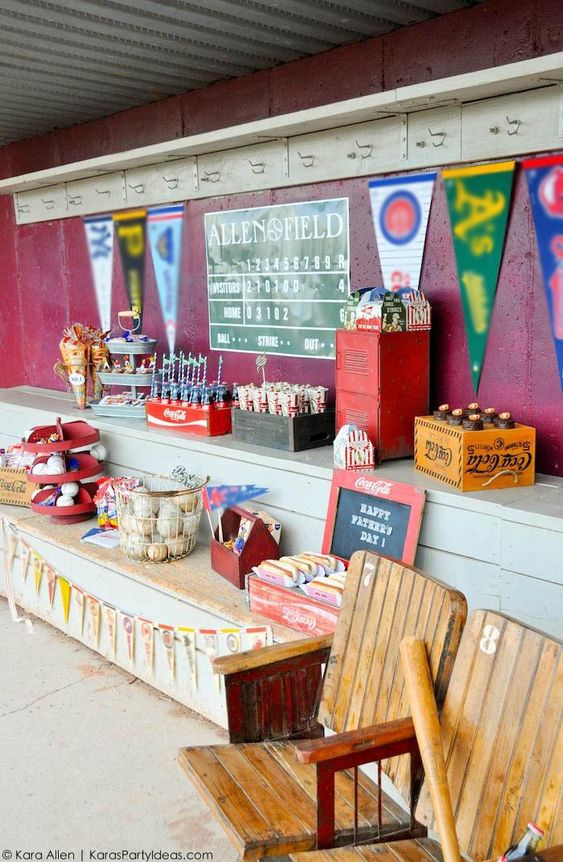 A baseball dugout decorated with old baseballs in wire baskets, wooden crates with sodas, popcorn, hot dogs, and treats.