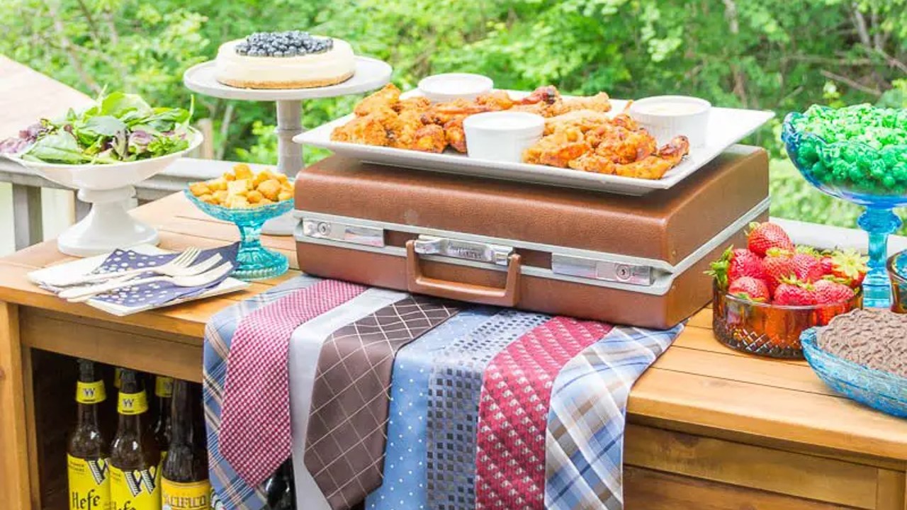 Buffet table set with a vintage briefcase set on a grouping of old men's ties. A platter of fried chicken strips sits on the briefcase. Cake plates and compotes surround the centerpiece and are filled with fruit, salads, and desserts. 