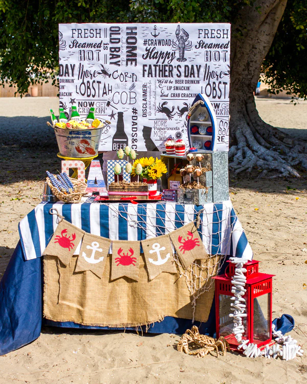 Outdoor dessert table backed by signage stating Happy Father's Day and lobster bake wording, along with a galvanized bucket holding beer bottle-shaped cookies, a sign that says DAD, sunflowers, and cake pops.