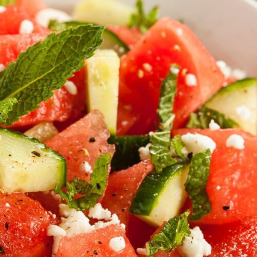 Cucumber watermelon salad garnished with mint leaves and topped with feta cheese crumbles in a white bowl.