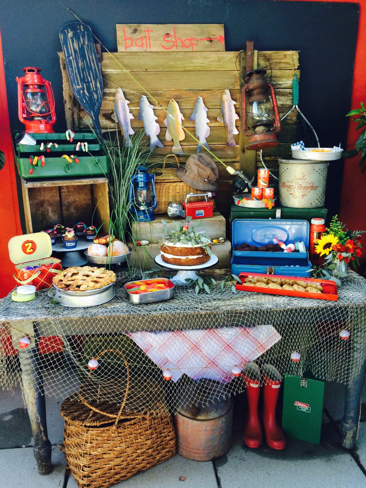 Dessert table decorated with a fishing net, tackle boxes, bobber, lanterns, fishing creels, fishing poles and wooden fish carvings.