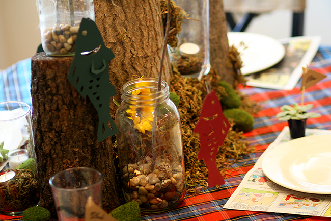 Tablescape featuring a centerpiece of three logs with mason jars holding tiny fishing poles made from twigs and twine. Paper fish are suspended from the fishing poles.