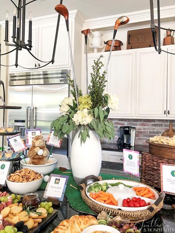 A golf-themed buffet featuring a floral arrangement that includes golf clubs, a golf green made from artificial turf that serves as the table runner, printed quotes from the Caddyshack movie, and finger foods. 