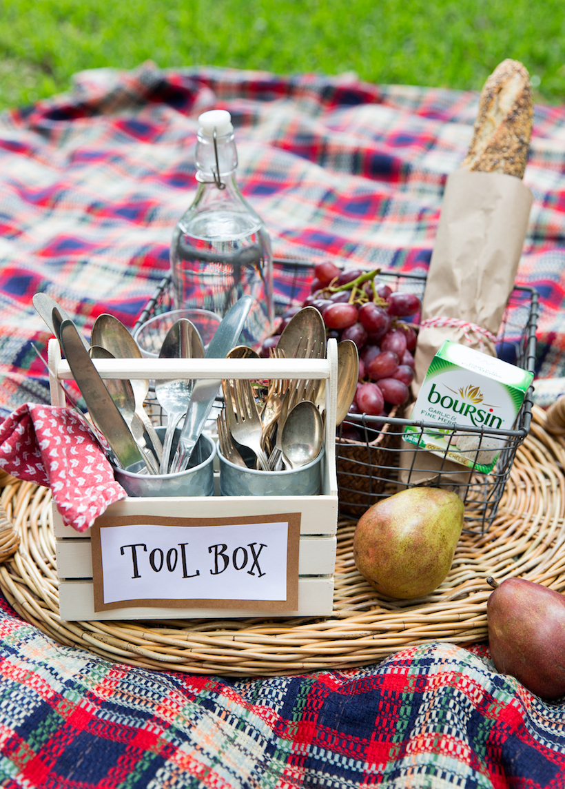 A picnic spread with a plaid blanket, fruit, bread, cheese and a silverware caddy with a sign that says "tool box."