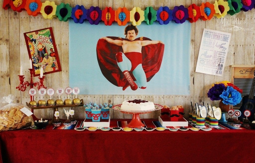 A Nacho Libre-themed buffet table with a red table cloth, poster of Jack Black in his Nacho Libre wrestling costume, red cake stand with a white frosted cake, treat bags with the wrestling mask on them, cookies, drinks, and chips.