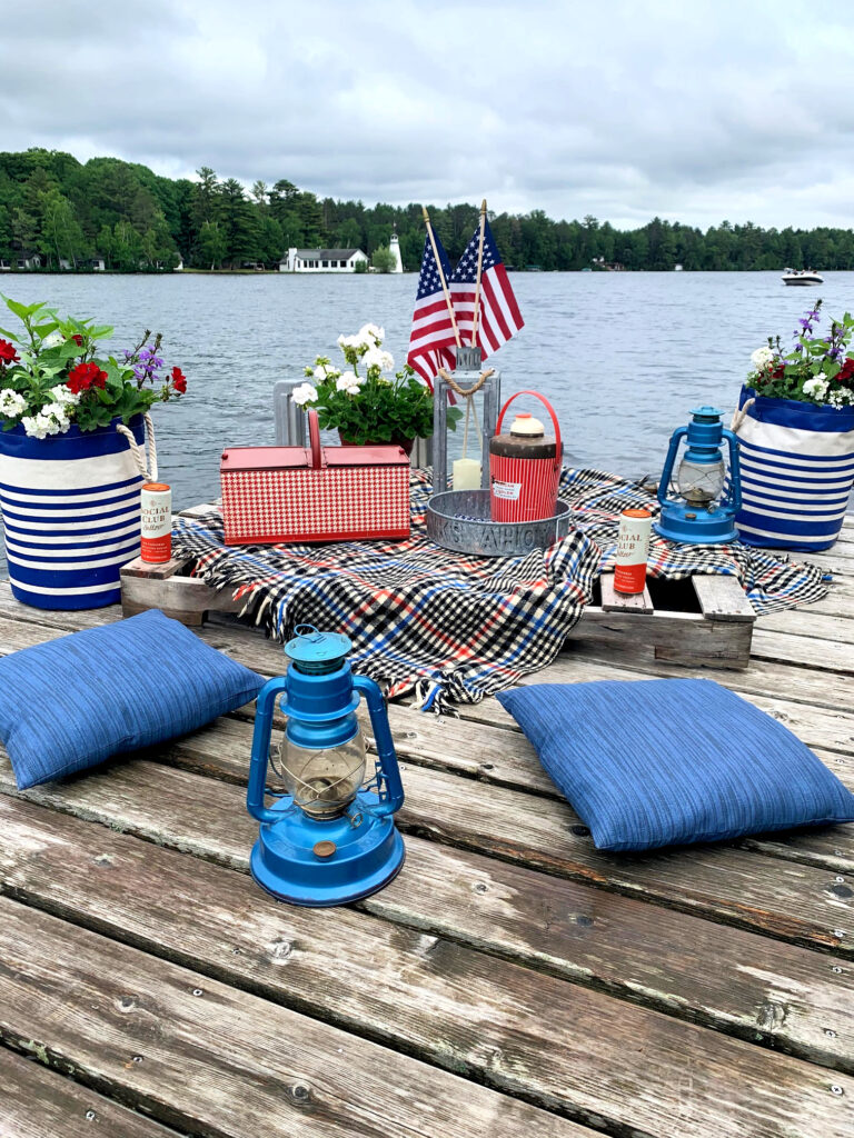 Picnic set up on a dock. Blue cushions, blue lanterns, a vintage red metal picnic basked, plaid blanket, and flowers.