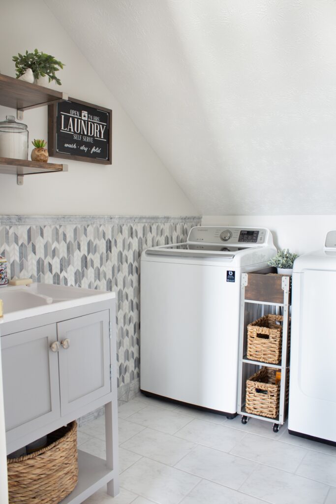 Laundry room featuring a narrow rolling cart parked between a white washer and dryer. The cart is a metal frame with wood shelves The top shelf features a wood box and the two lower shelves hold baskets. 