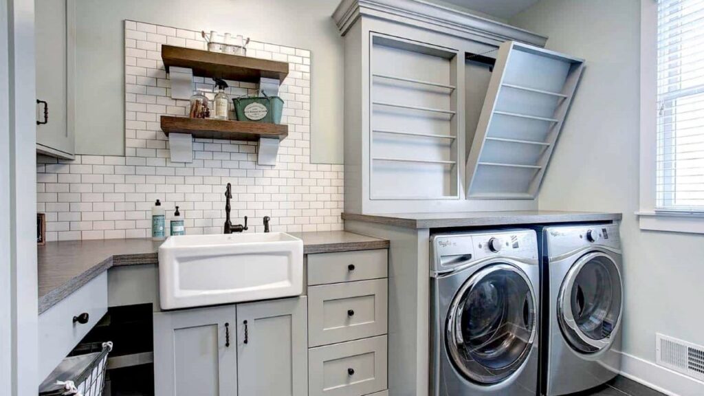 Laundry room in light gray tones with a white apron-front sink, wood counters, gray painted cabinets, and stainless steel front load washer dryer.