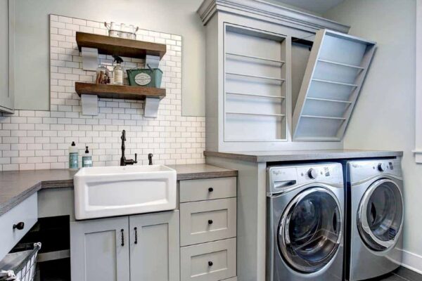 Laundry room in light gray tones with a white apron-front sink, wood counters, gray painted cabinets, and stainless steel front load washer dryer.