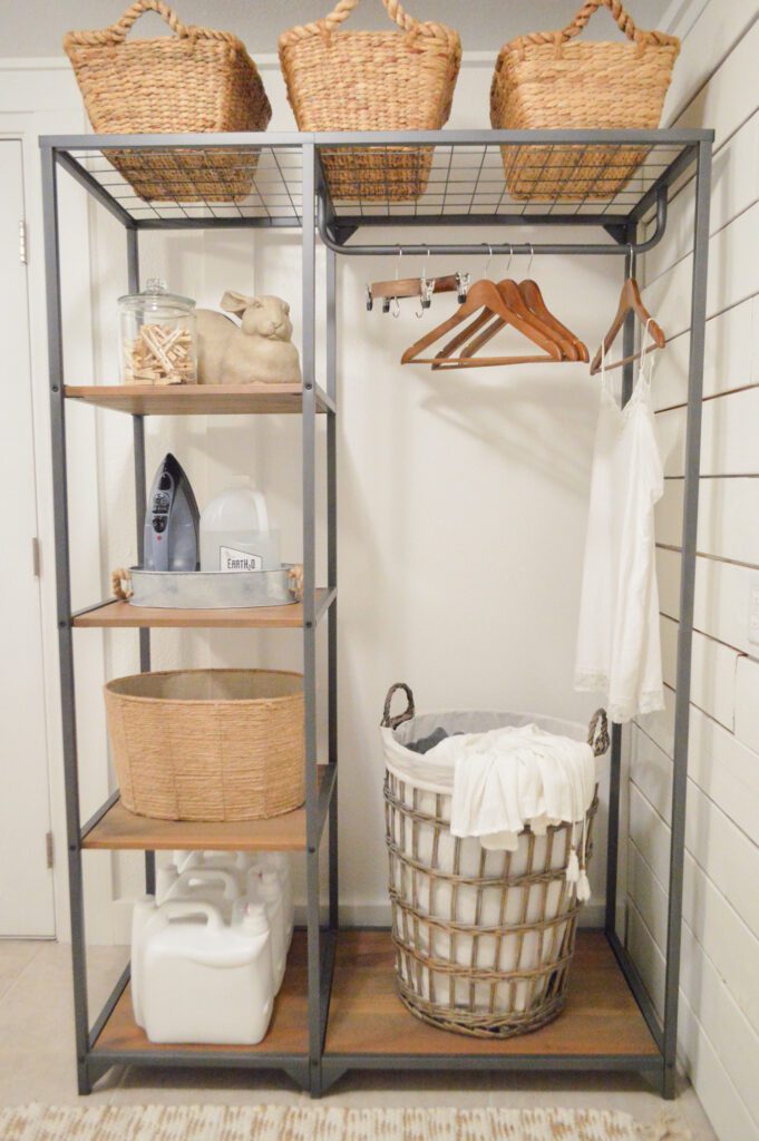 Metal framed stand with wood shelves on the left-hand half holding baskets and supplies. The right-hand side features a hanging rod for clothes and a bottom wooden shelf holding a laundry basket.