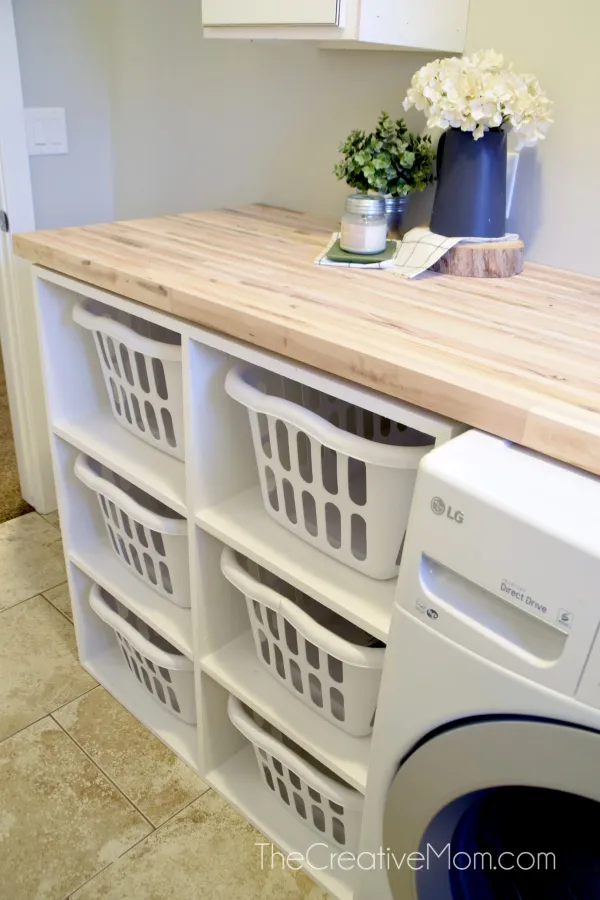 A DIY laundry basket organizer. It's six cubbies made from white laminate and is stored under a wooden countertop.