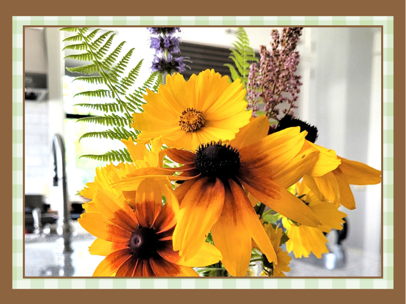 Wildflowers-Closeup | The Granite Acorn Bright bouquet of orange and yellow flowers with dark centers and green ferns on a kitchen counter