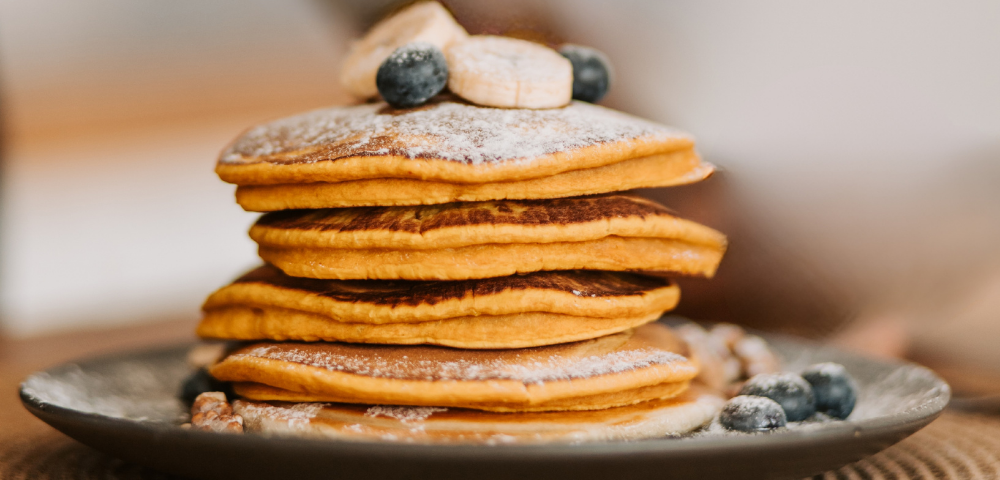 Stack of pumpkin pancakes on a plate with sliced bananas, blueberries, and powdered sugar on top.