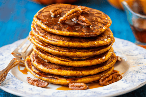 Stack of pumpkin pancakes on a plate with maple syrup and pecans.