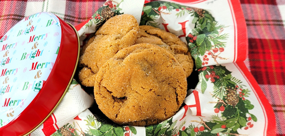 Metal tin lined with a red and green holiday napkin. The tin is filled with crinkle top cookies and it sits on a red and green plaid cloth.