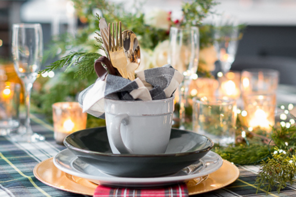 Table setting with blue plaid table cloth, red plaid napkins, blue and white dishes, votive candles, fairy lights and an evergreen centerpiece