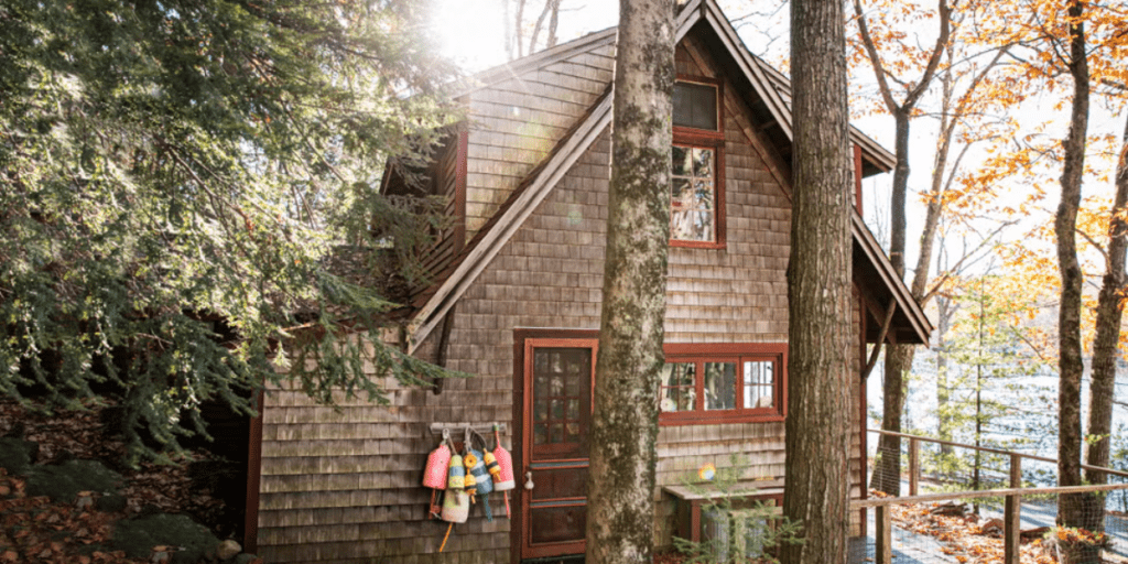 Shingle sided house with red trim surrounded by woods on the edge of a pond.