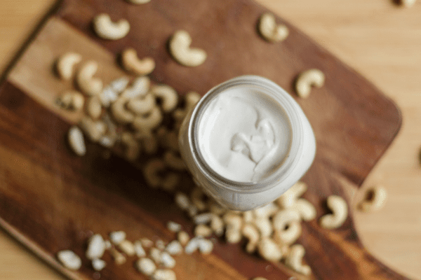 Cashew Cream-Banner | The Granite Acorn Canning jar filled with cashew cream on a wooden cutting board scattered with raw cashew nuts