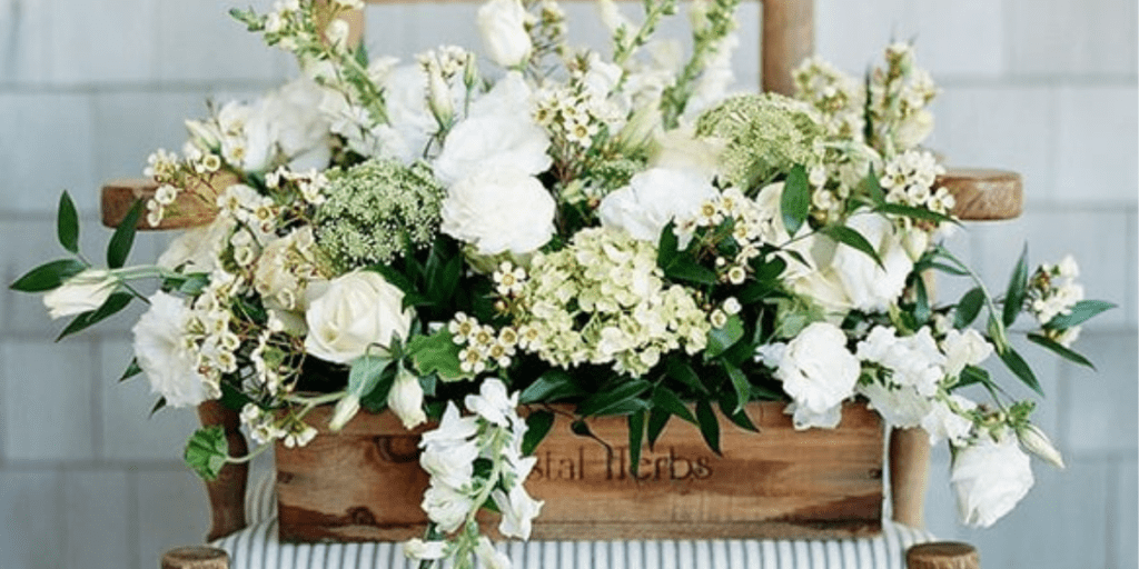 Low wooden box filled with white hydrangea, roses, and ranunculus sitting on a ladderback chair with a blue and white striped fabric seat.