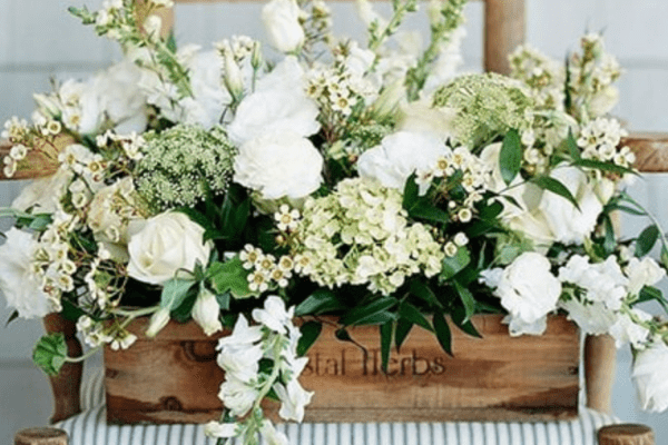 Low wooden box filled with white hydrangea, roses, and ranunculus sitting on a ladderback chair with a blue and white striped fabric seat.