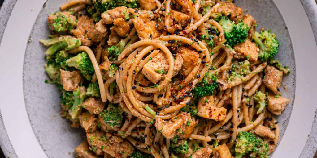 Closeup of serving bowl with whole wheat noodles, broccoli, and tofu chunks in an almond butter sauce.