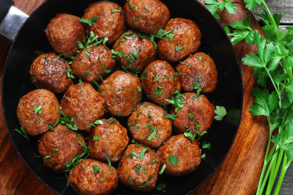Vegan Meatballs-banner | The Granite Acorn Skillet filled with vegan meatballs sprinkled with chopped parsley on a wooden cutting board Sprigs of fresh parsley are laid to the side of the skillet