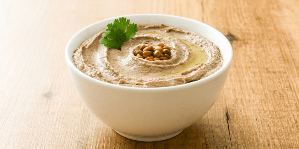 Small white bowl of hummus on a wooden counter. The hummus is garnished with parsley, olive oil, and crispy chickpeas.