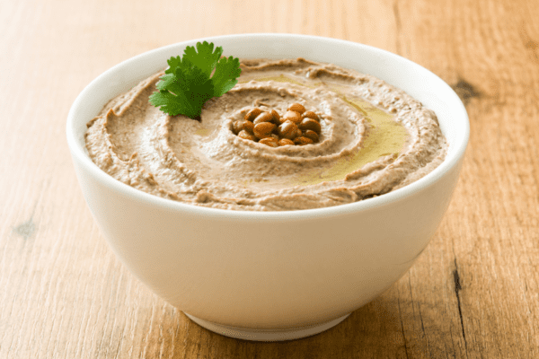 hummus-banner | The Granite Acorn Small white bowl of hummus on a wooden counter The hummus is garnished with parsley olive oil and crispy chickpeas