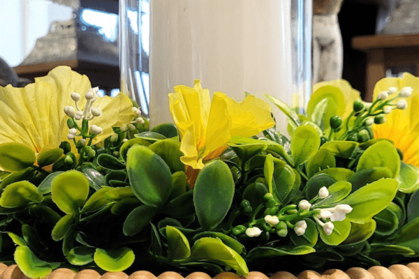 Spring-Candle-Ring-Banner | The Granite Acorn Close up of a boxwood candle ring with yellow flowers and white seed heads There's a white pillar candle in the center in a glass vase and it's sitting on a round wood stand