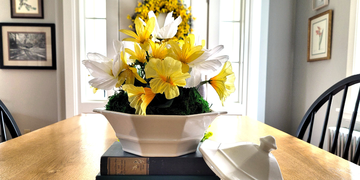 Yellow and white floral centerpiece in a white ceramic vegetable bowl on a stack of green books