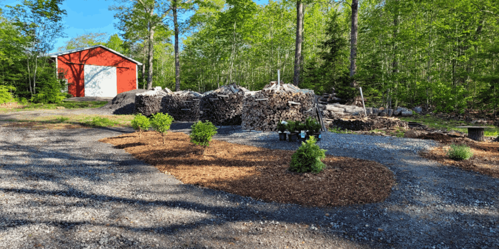 Spring to Fall 2025 | The Granite Acorn Round wood piles bordered by gravel paths and newly planted hydrangea with a red barn in the background