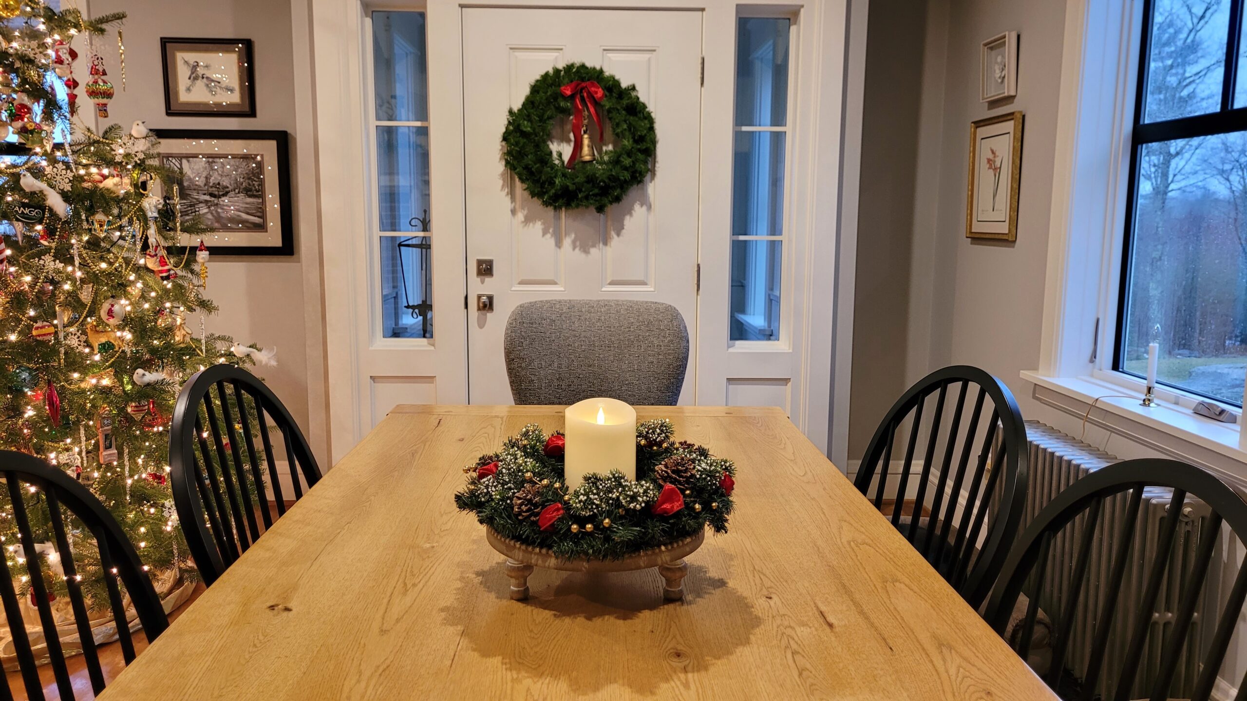 Oak dining room table with a small wreath candle ring around an ivory pillar candle Another wreath hangs on a door in the background
