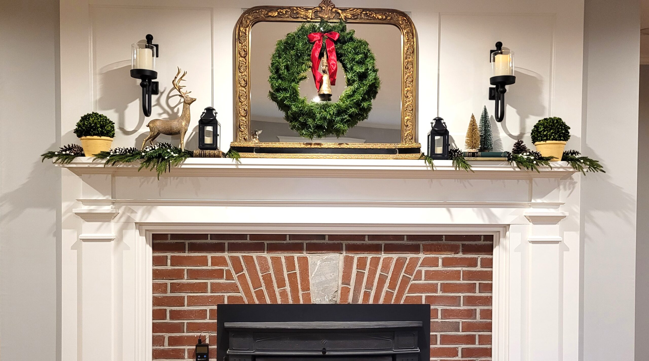 Red brick fireplace with a white painted wood mantle A large gold mirror with a christmas wreath is on the mantle along with pine boughs preserved boxwood balls a gold deer small lanterns and bottle brush trees