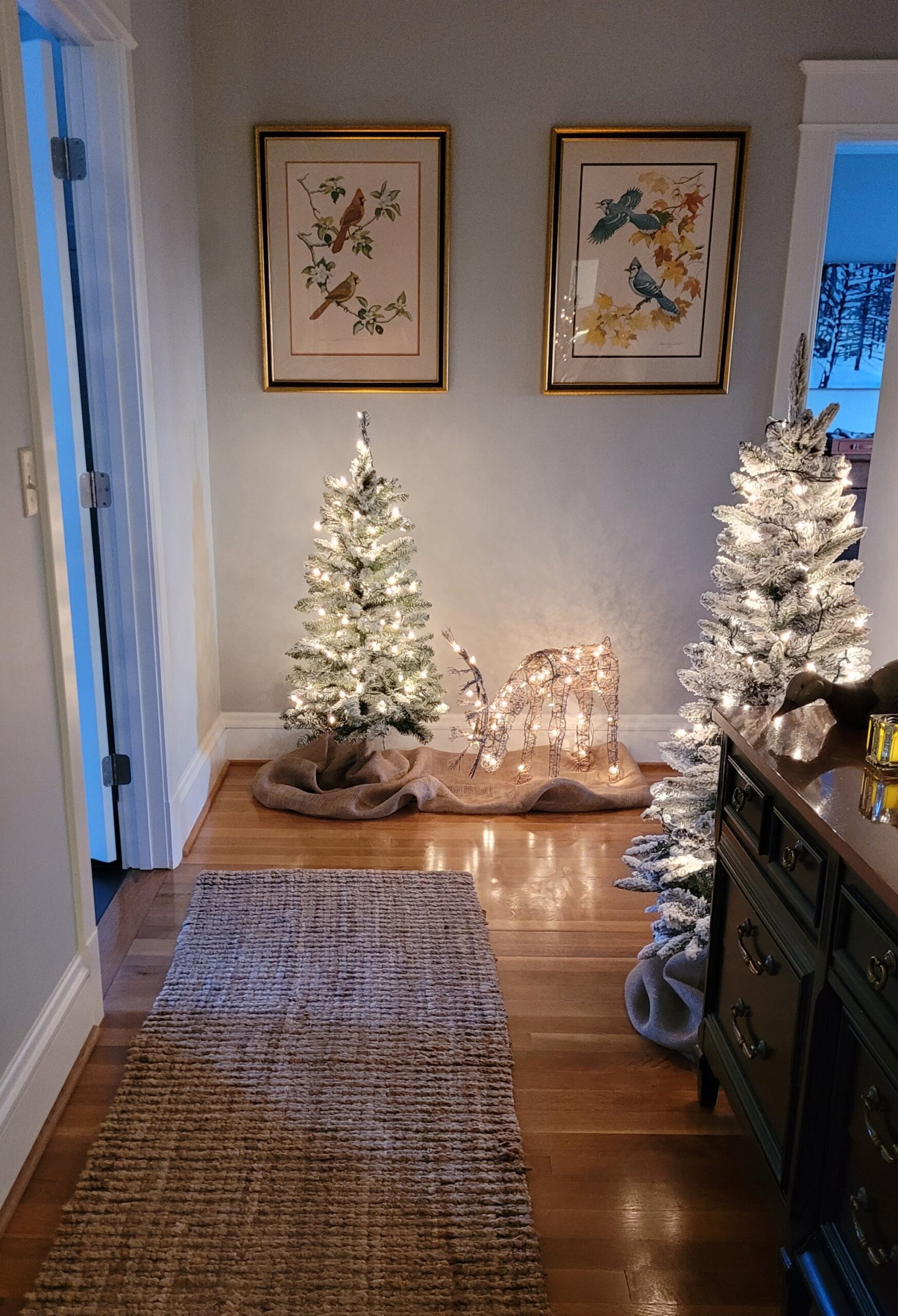 Photo of hallway featuring a 4 foot flocked tree that is lit up and a grapevine deer that is lit up The tree and deer are on a length of burlap