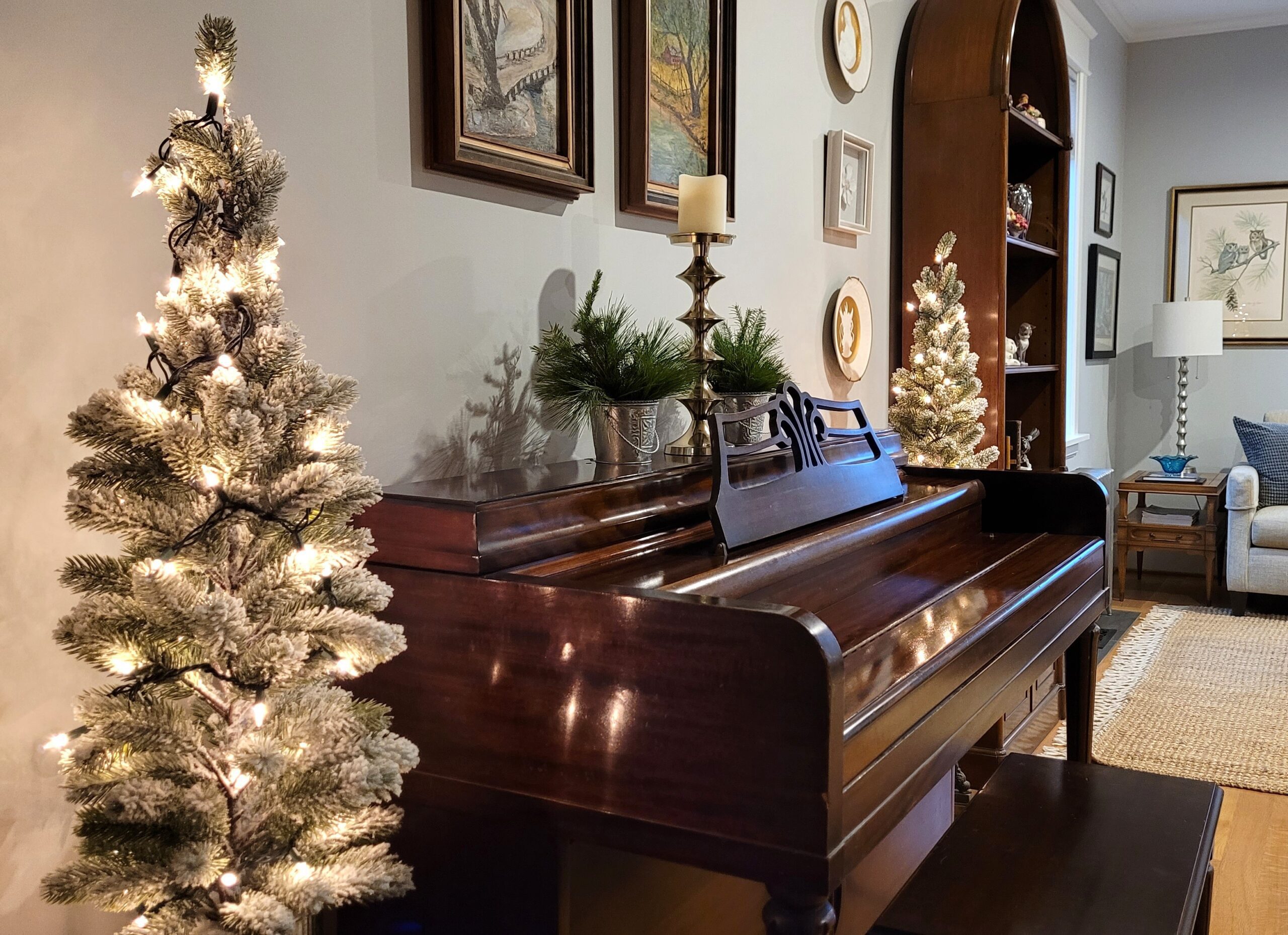 Angled photo of a mahogany wood piano flanked by two flocked and lit trees on round wood end tables On the piano is a gold candlestick and two metal pails with fresh evergreen sprigs