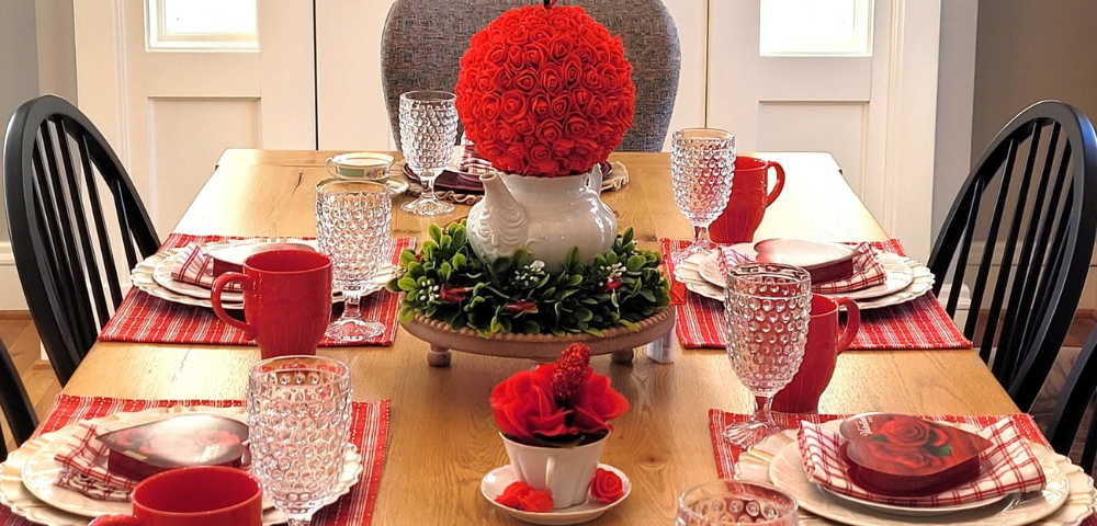 Wood dining table set with red placemats, white china, and a red rose covered topiary in the middle.