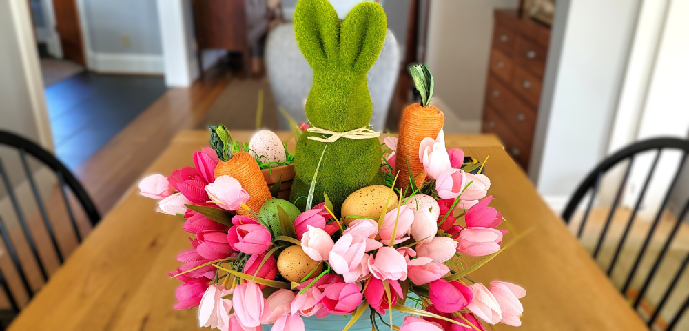 Easter centerpiece featuring a green flocked bunny in a light blue pail surrounded by pink tulips, jute carrots, and faux Easter eggs.