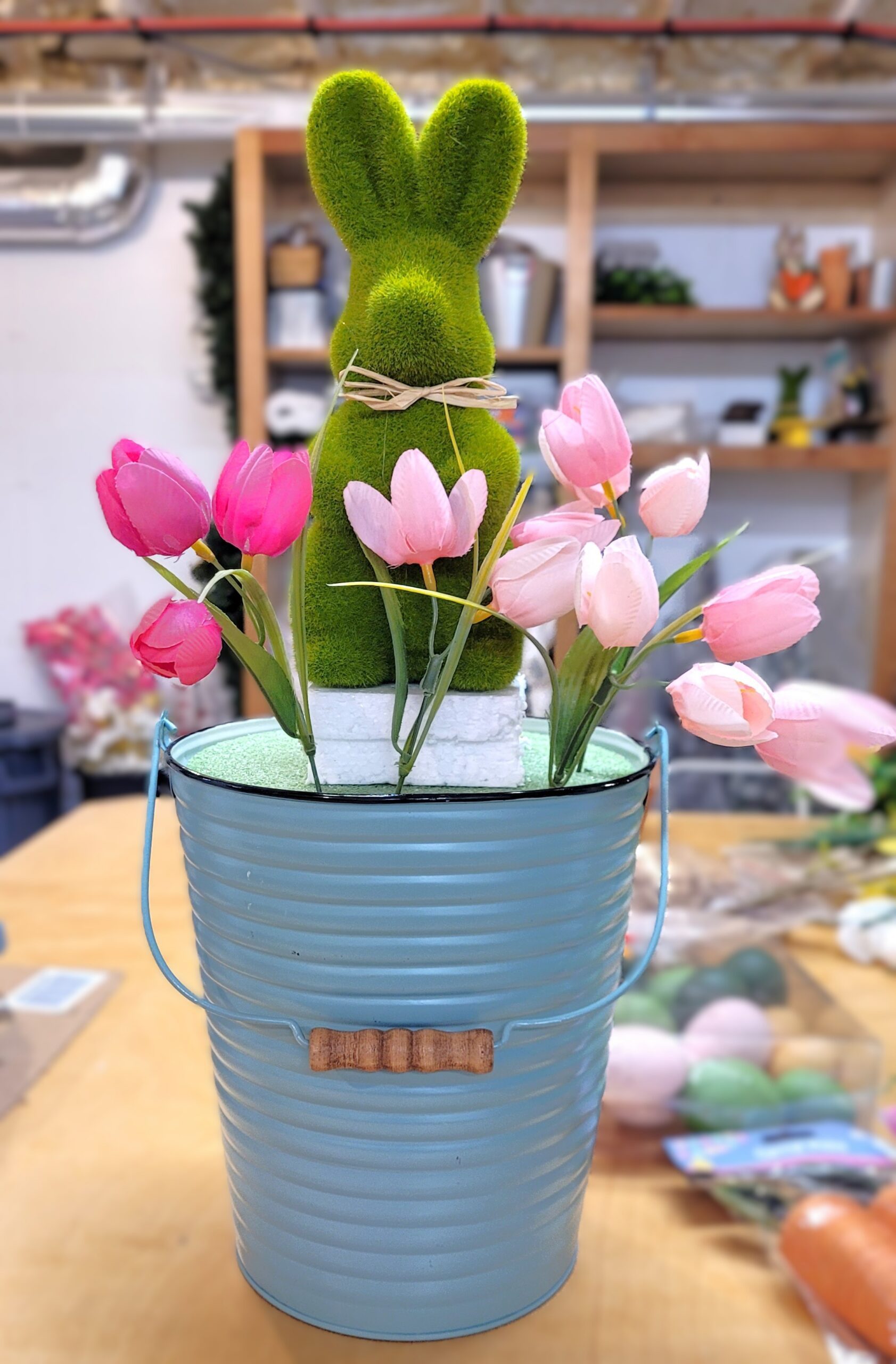 Green moss bunny centerpiece being made The bunny is attached to foam blocks in a pastel blue pail Pink tulips have just begun to be added