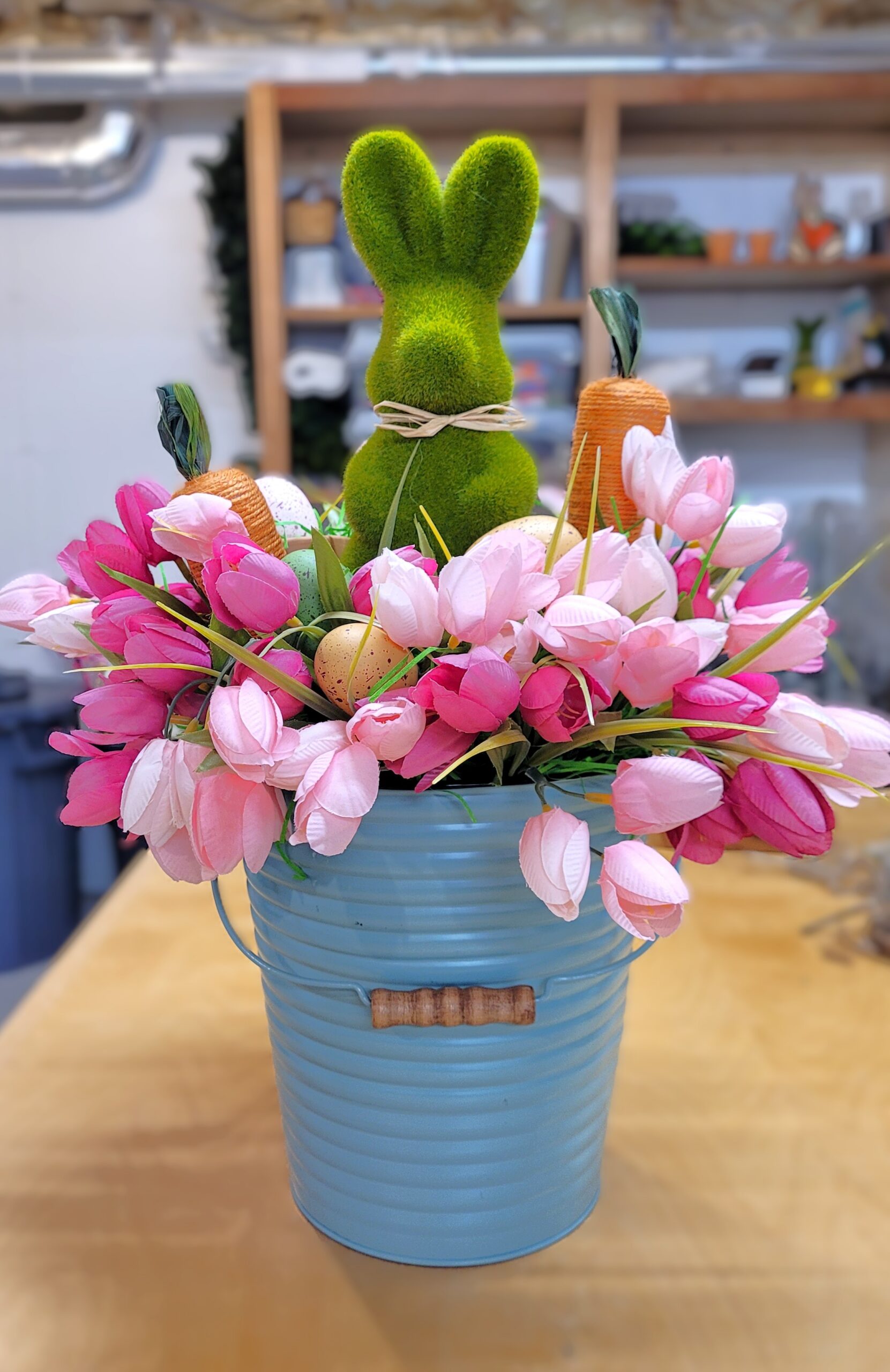 Green moss bunny surrounded by pink tulips faux easter eggs and jute carrots The arrangement is in a pastel blue metal pail