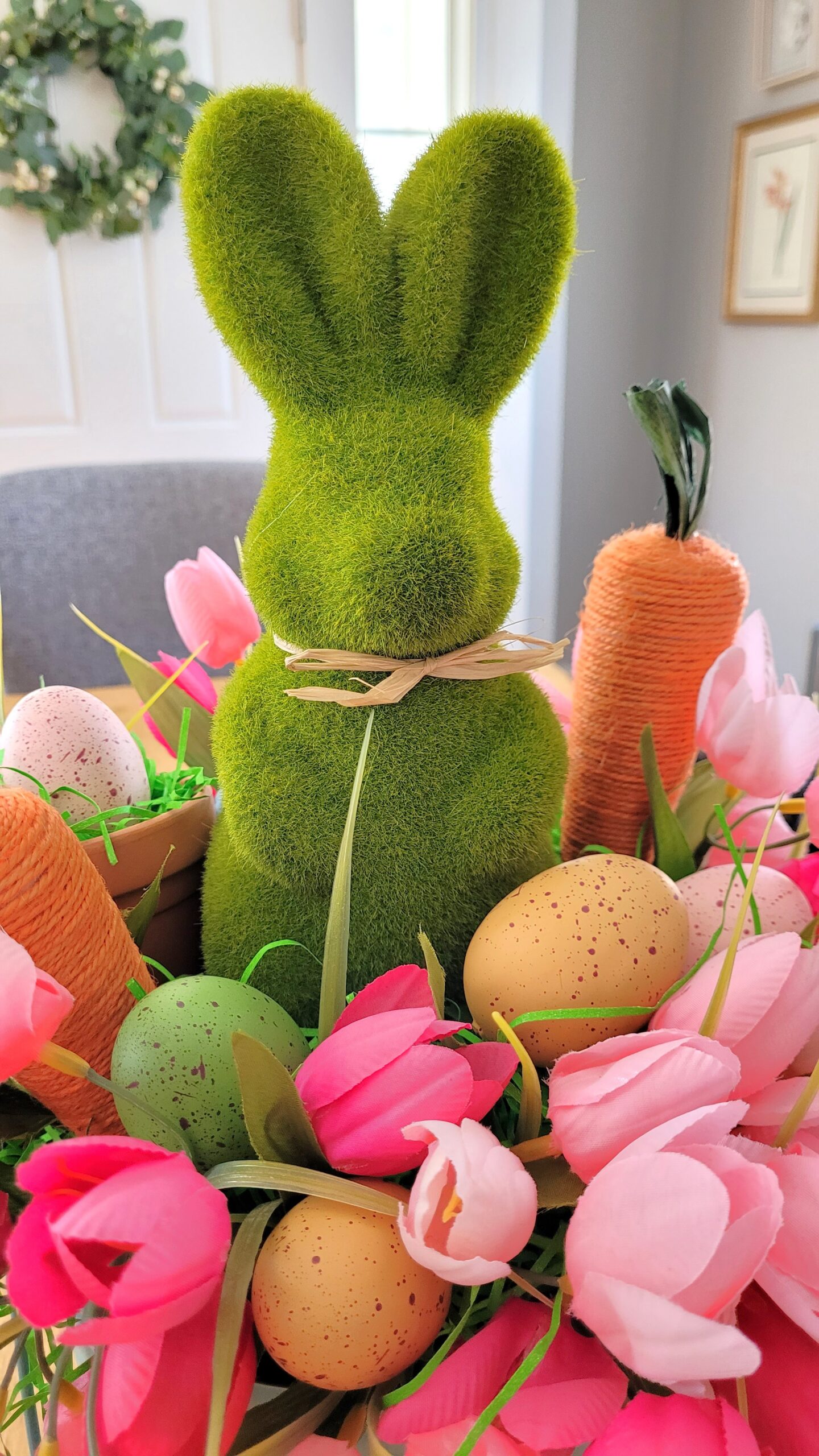Closeup of a green moss bunny surrounded by pink tulips faux easter eggs and jute carrots